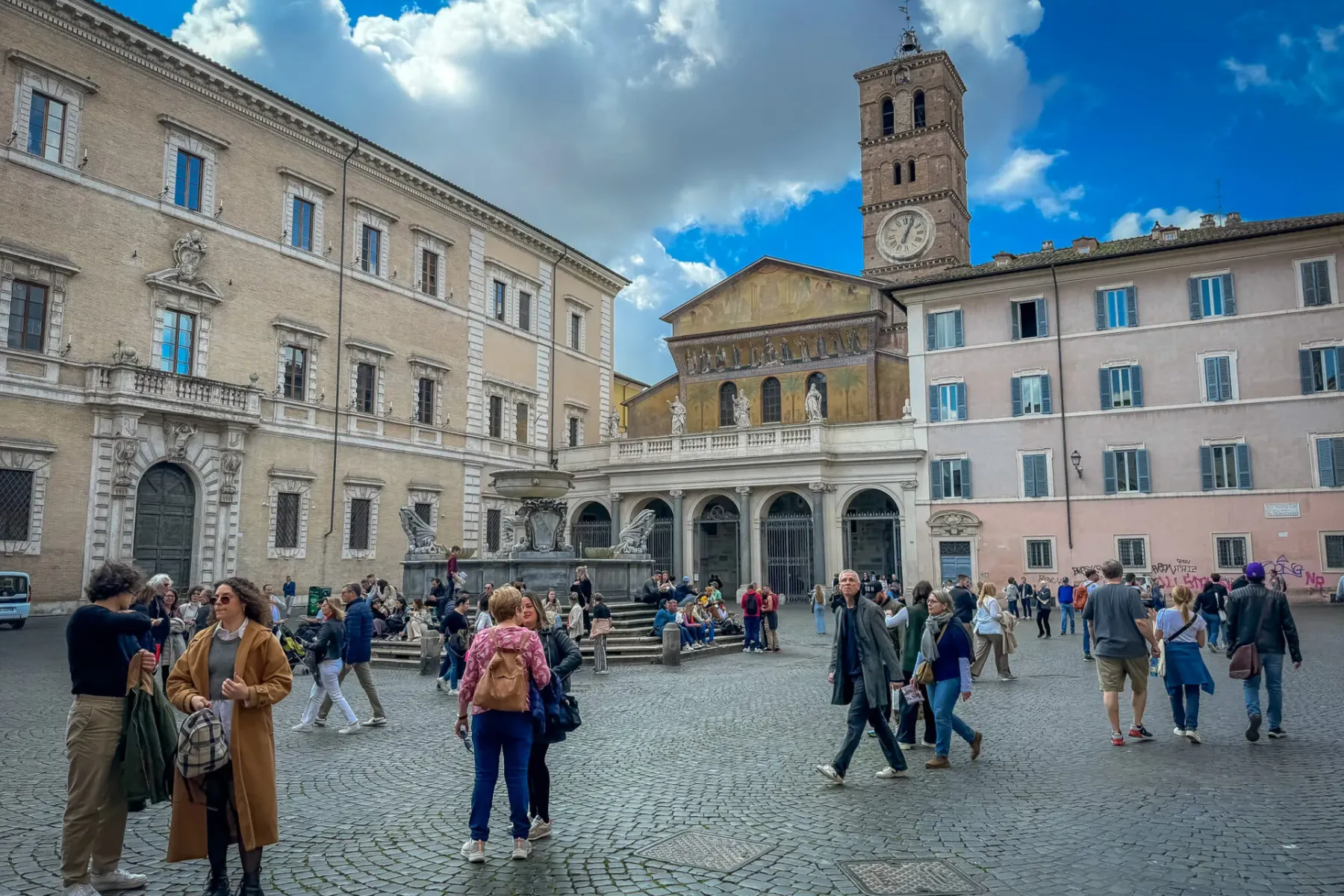 Adoquinada Piazza Santa Maria en Trastevere, Roma, con basílica, campanario, fuente y muchas personas paseando.
