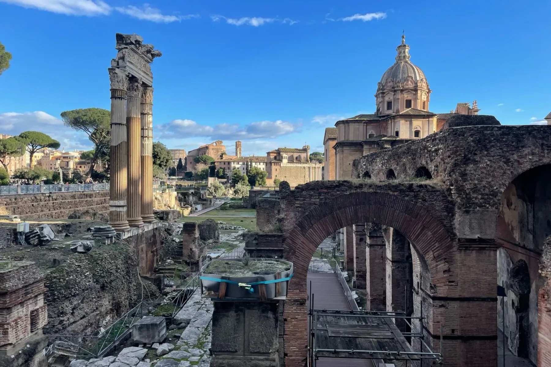 Ruinen des Forum Romanum mit Säulen des Saturn-Tempels, weiteren Tempeln und Besuchern unter blauem Himmel.