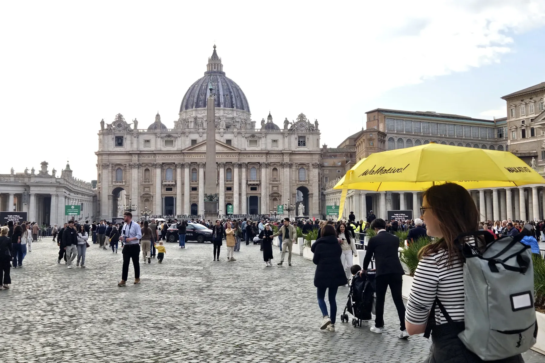 Belebter Petersplatz mit dem Petersdom, einem Obelisken und Kolonnaden unter teilweisem Himmel. Ein gelber Walkative-Regen...