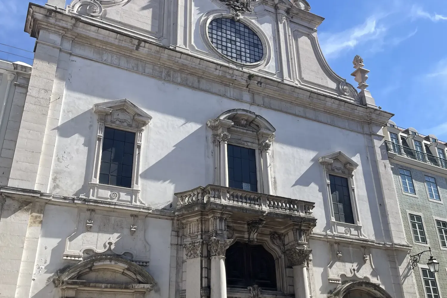 Verzierte weiße Fassade der São Domingos Kirche in Lissabon, mit großen dunklen Türen, einem Balkon und einem Kreuz auf de...