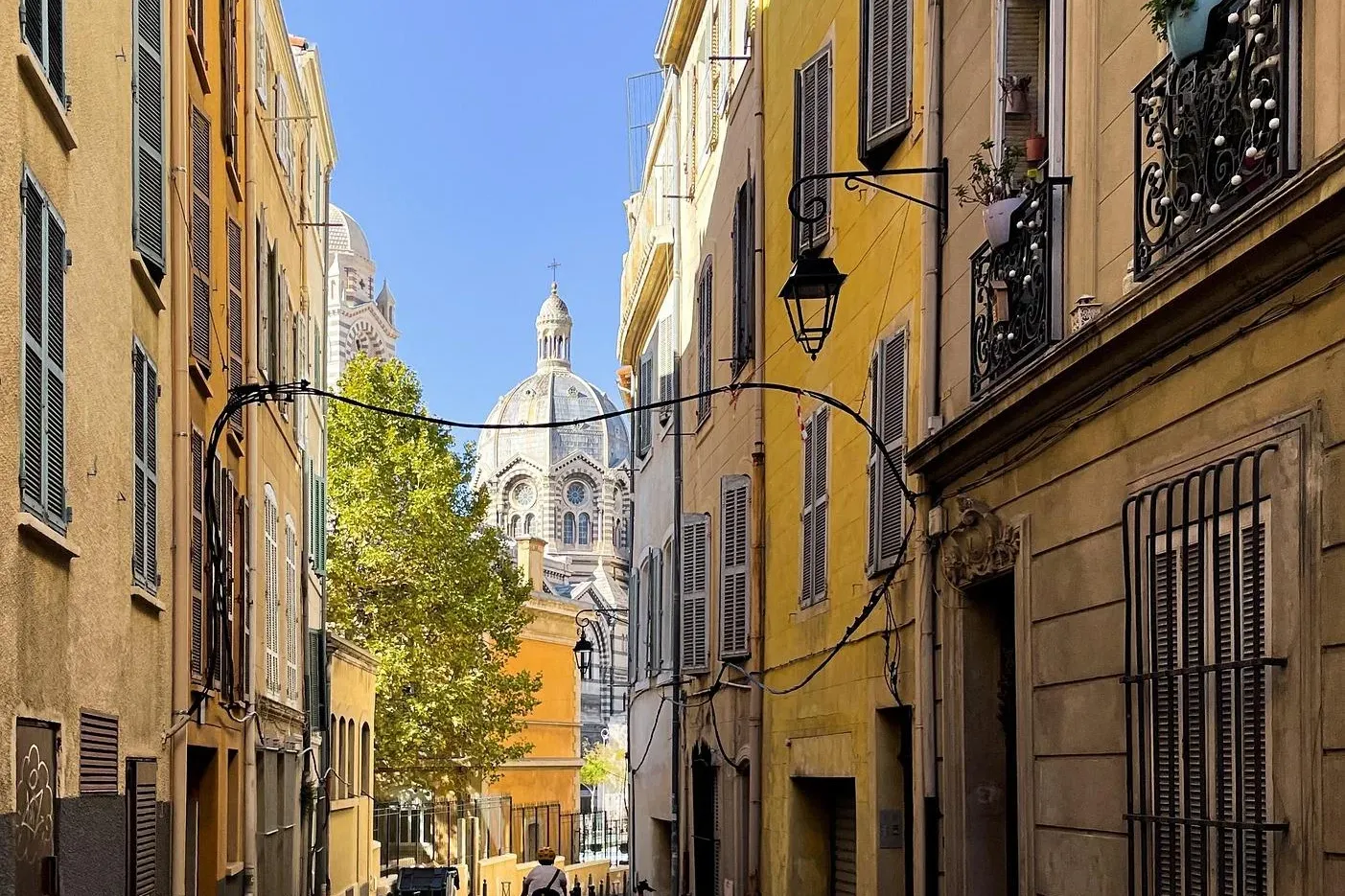 Calle estrecha en Marsella flanqueada por edificios amarillos con persianas, que conduce a la cúpula de Notre-Dame de la G...