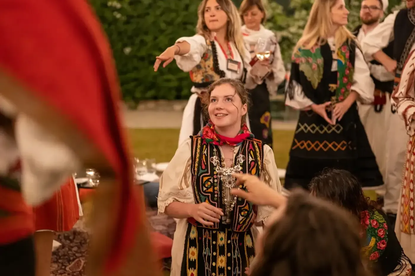 Młoda kobieta w haftowanym tradycyjnym stroju albańskim i czerwonej chuście uśmiecha się, z innymi osobami w tle.