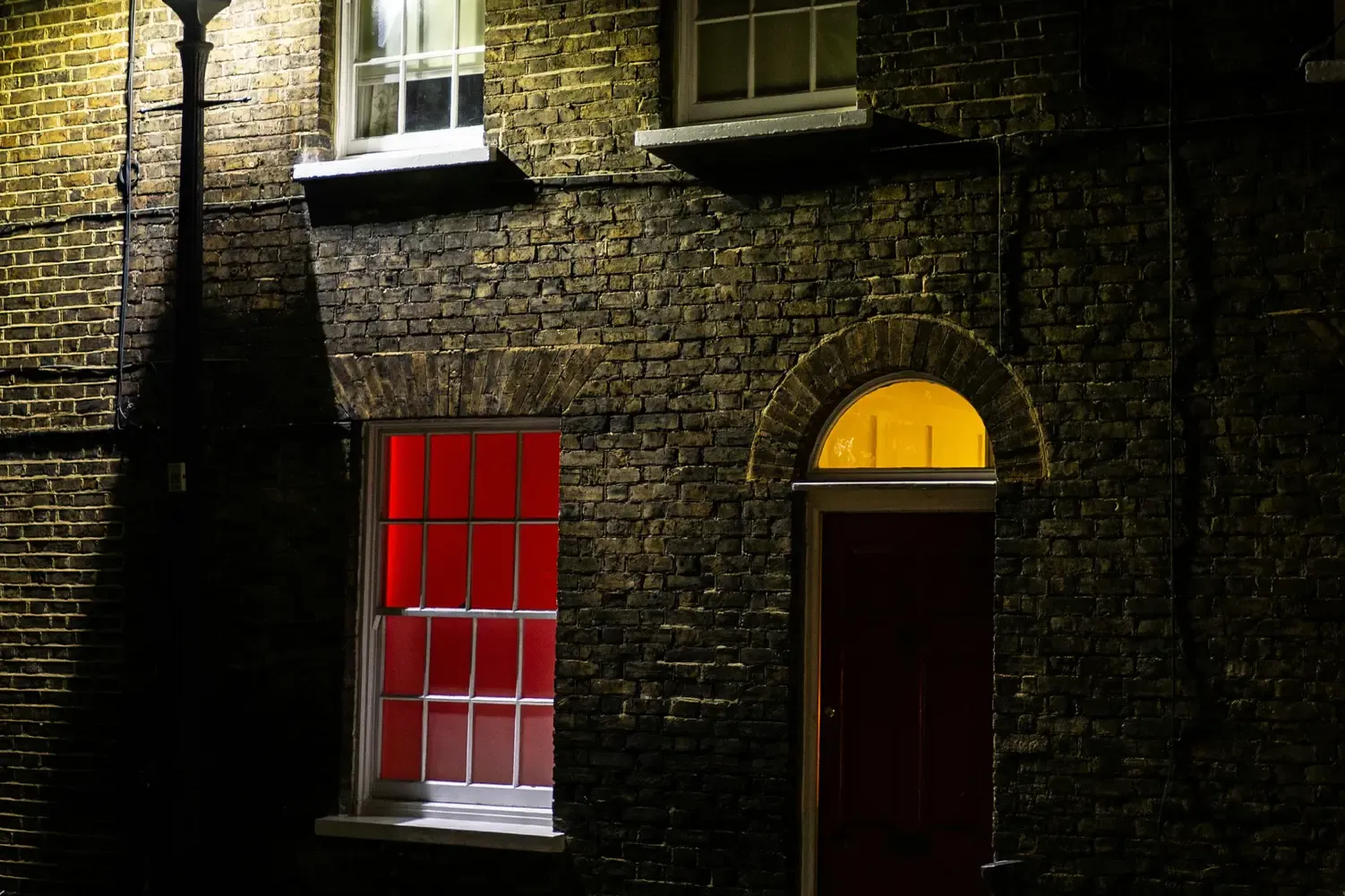 Edificio de ladrillo en una calle oscura de noche, iluminado por una farola. Una ventana brilla con una luz roja.