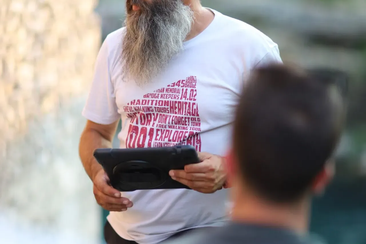 Brodaty przewodnik z okularami na głowie trzyma tablet, rozmawiając z grupą na zewnątrz w Mostarze.