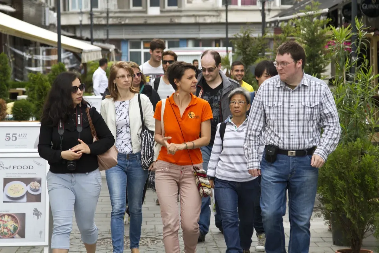 Un grupo diverso de personas, incluyendo un guía con polo naranja, camina por una calle pavimentada en Bucarest, pasando e...