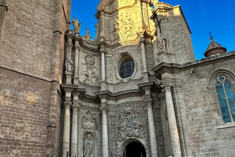 Detailed baroque facade of Valencia Cathedral with a prominent tower and entrance, bathed in sunlight against a deep blue ...