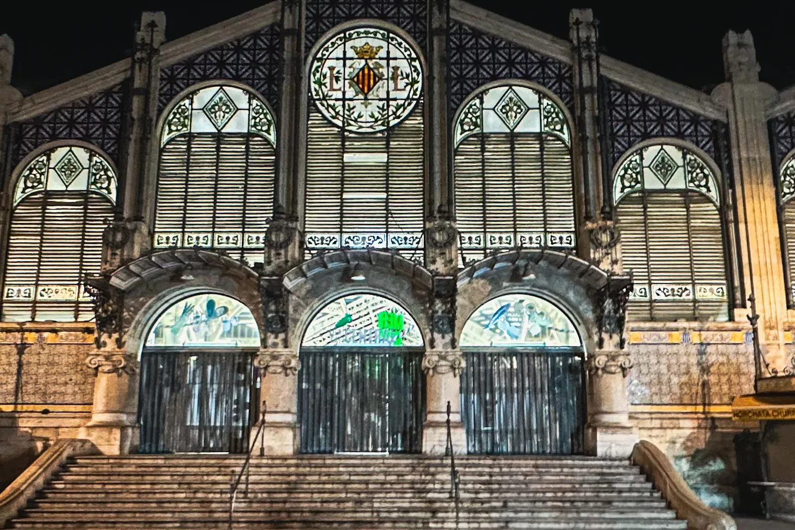Illuminated Art Nouveau facade of Valencia Central Market at night, featuring arched entrances, steps, and intricate stain...
