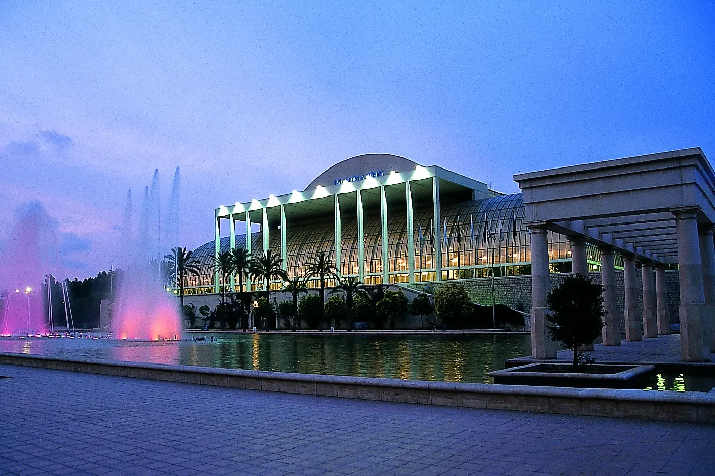 The Palau de la Música in Valencia at dusk, its glass facade lit up, with colourful fountains in the foreground.