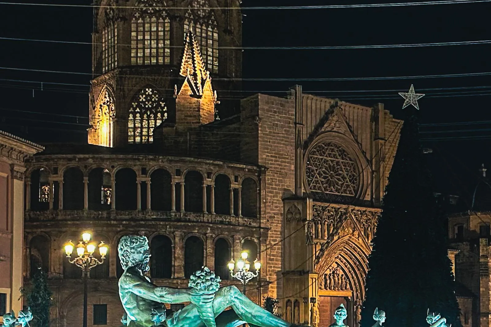 Illuminated Turia Fountain with bronze statues and the illuminated Valencia Cathedral at night. A large Christmas tree is ...