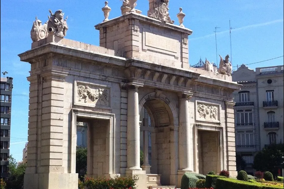 The Puerta del Mar triumphal arch in Valencia on a sunny day, with a bed of red flowers in the foreground.