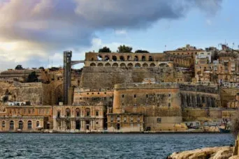Vista de las históricas fortificaciones y edificios de piedra de La Valeta a lo largo del Gran Puerto, bajo un cielo nublado.