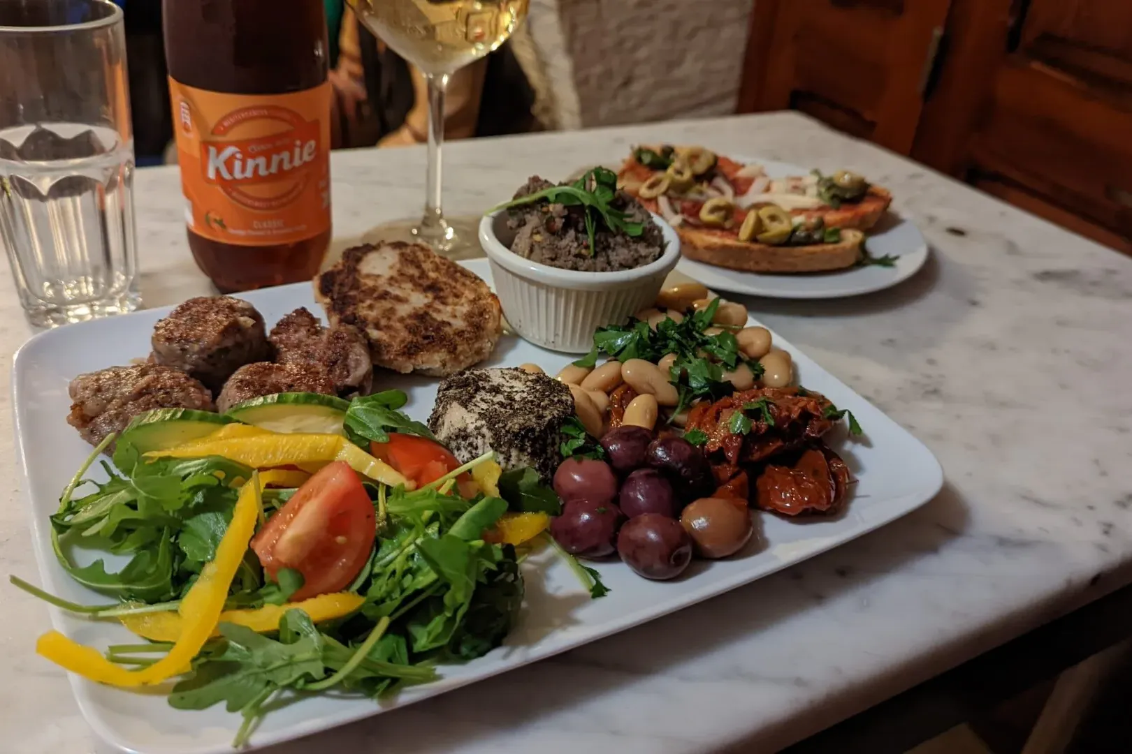 Eine Platte maltesischer Speisen mit Frikadellen, Salat, Bohnen, Oliven und Käse, dazu eine Kinnie-Flasche und Weinglas au...