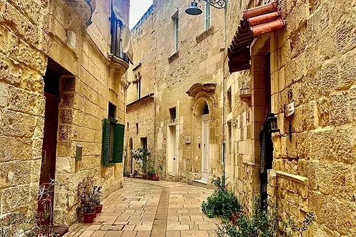 A narrow stone street in an old town, flanked by tall buildings with balconies, green shutters, and ornate street lamps.