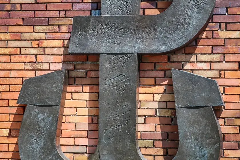 Duża, ciemna metalowa rzeźba symbolu Polski Walczącej (Kotwicy) na wielobarwnym ceglanym murze z liśćmi u góry i bluszczem...