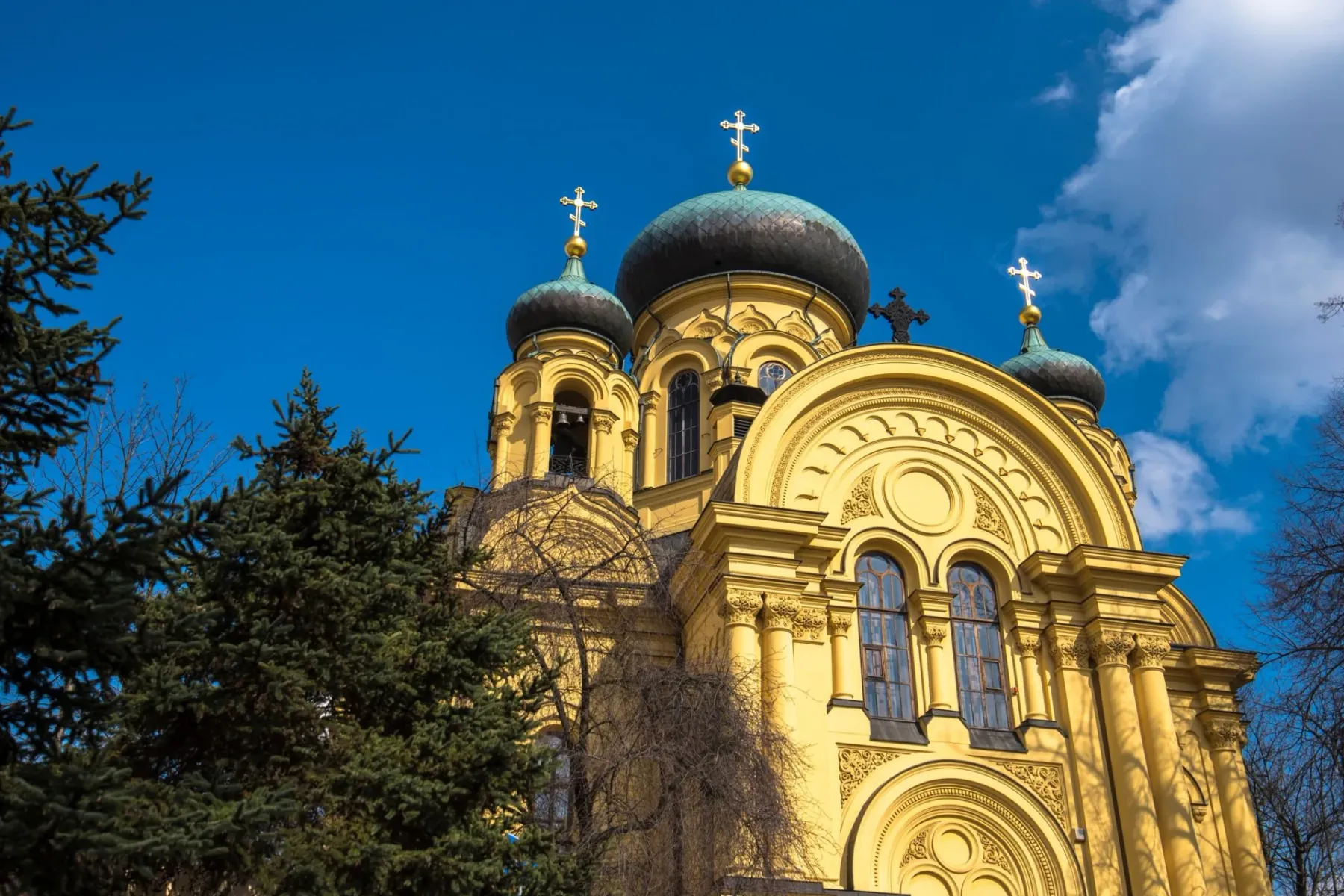 Yellow Orthodox church with dark domes and golden crosses against a blue sky, surrounded by green trees.