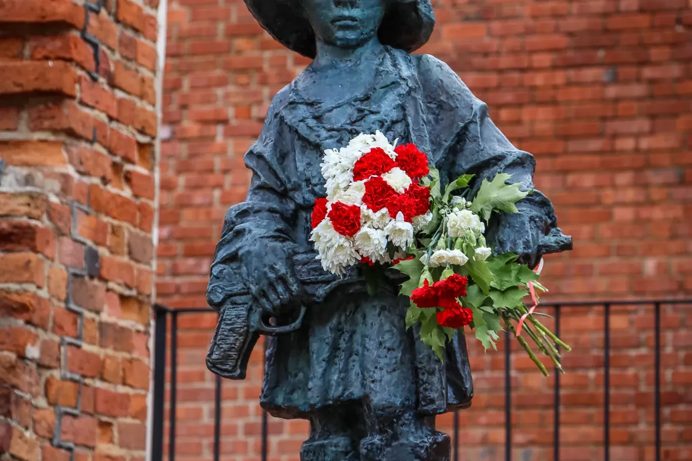 Brązowy pomnik małego powstańca w hełmie, trzymającego biało-czerwone kwiaty, na tle ceglanej ściany.