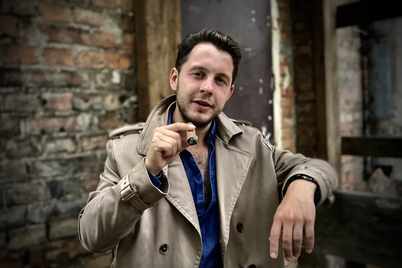 Young man in a beige trench coat and blue shirt holding a lit cigar, standing in front of an old brick wall.