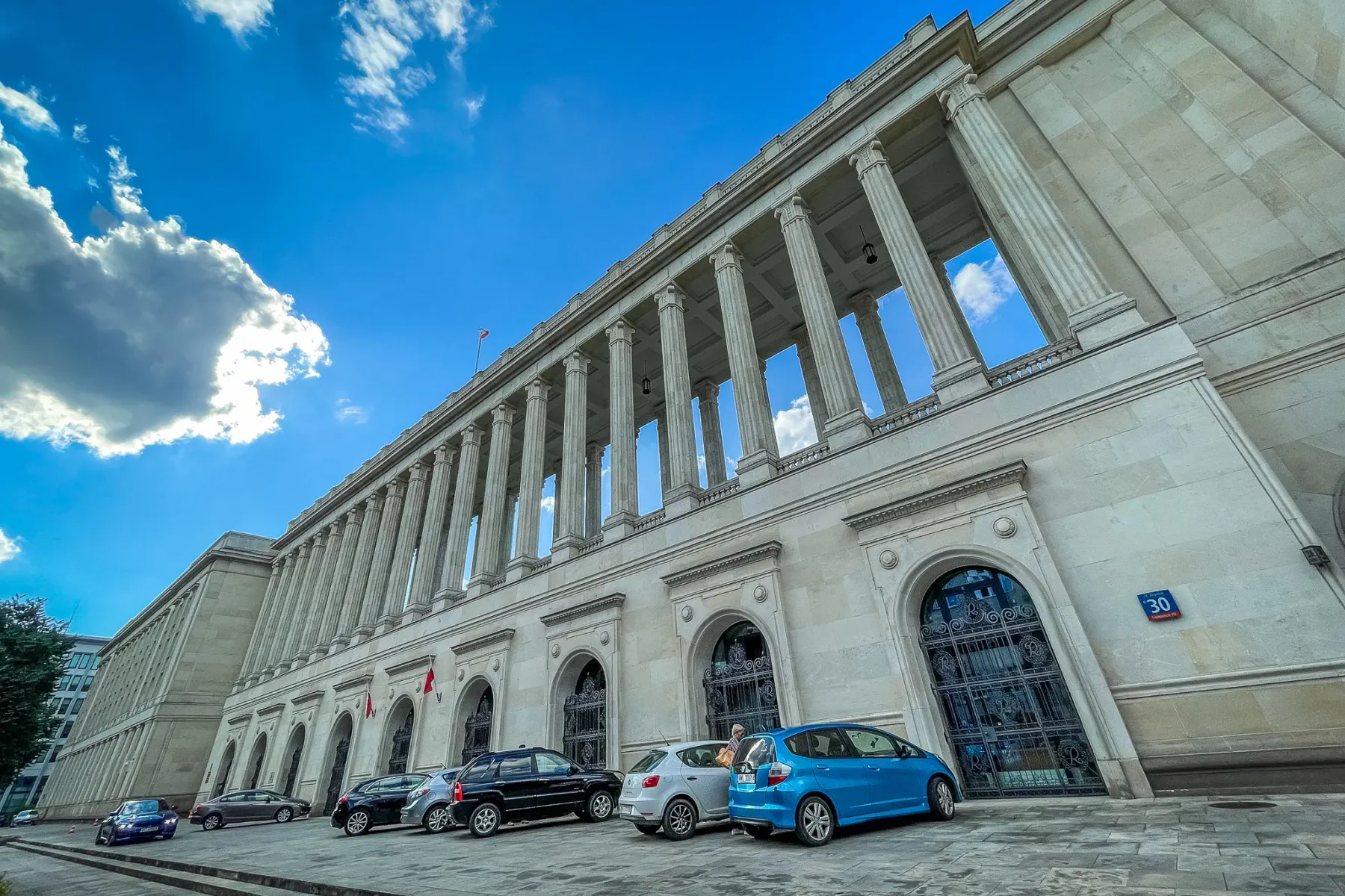 Large, imposing building with a long colonnade and parked cars under a bright blue sky in Warsaw.