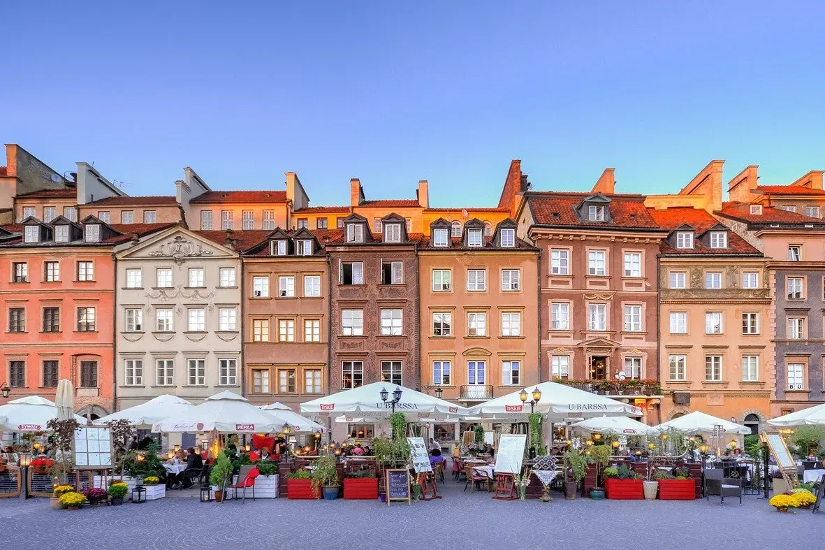 Farbige historische Gebäude mit roten Dächern überblicken einen Außenbereich mit weißen Sonnenschirmen und Menschen unter ...