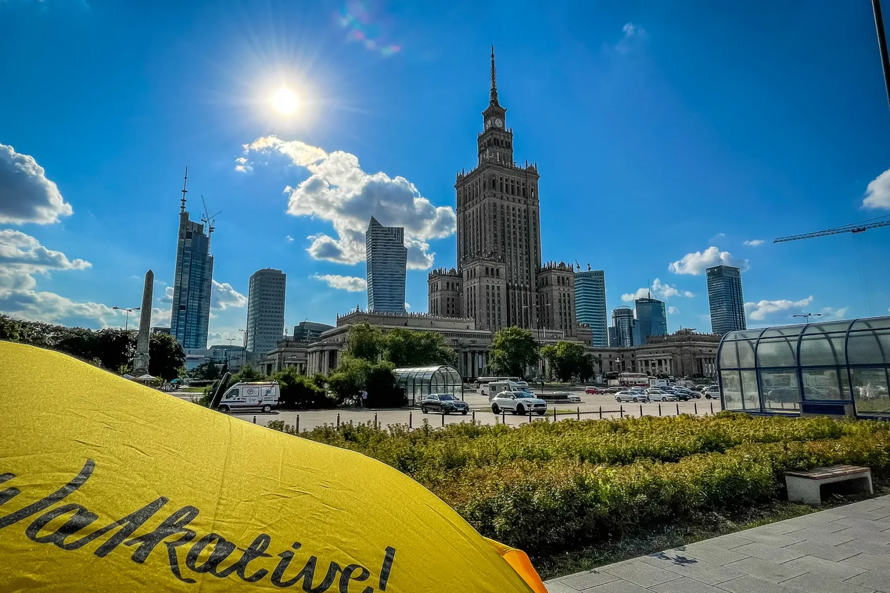 Palace of Culture and Science and modern skyscrapers under a sunny blue sky, with a yellow Walkative umbrella in the foreg...