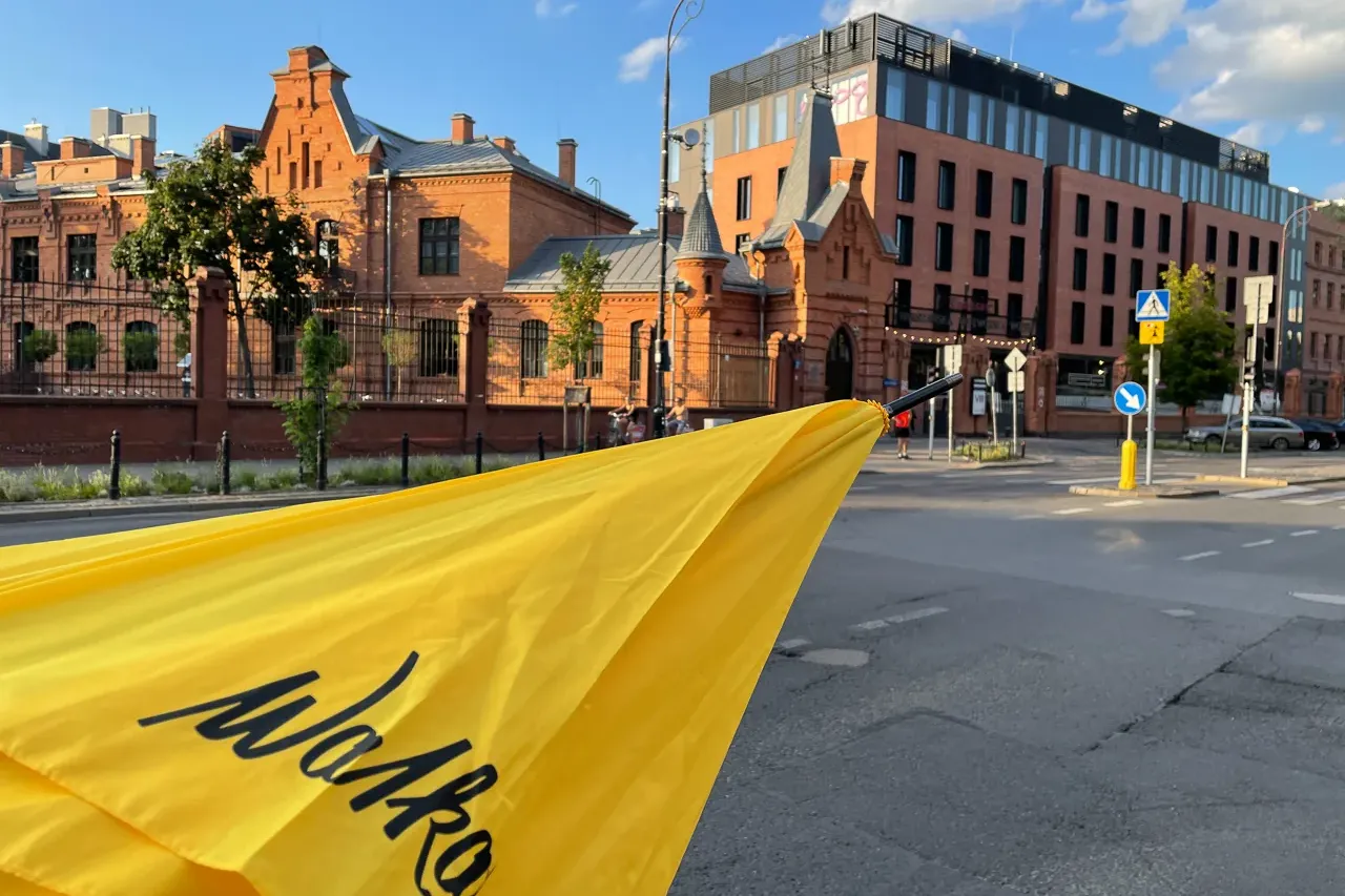 A yellow Walkative umbrella in the foreground, with historical brick buildings, a modern building, and a street in Warsaw'...