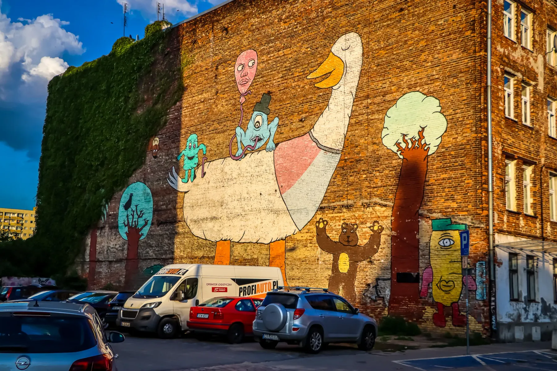 Colorful mural on a brick building in Warsaw, showing a large goose, whimsical figures, and a bear, with parked cars below.