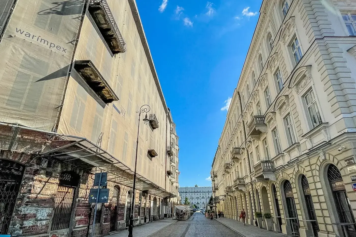 Cobblestone street between a scaffolded building with 'warimpex' and an ornate historical building with balconies under bl...