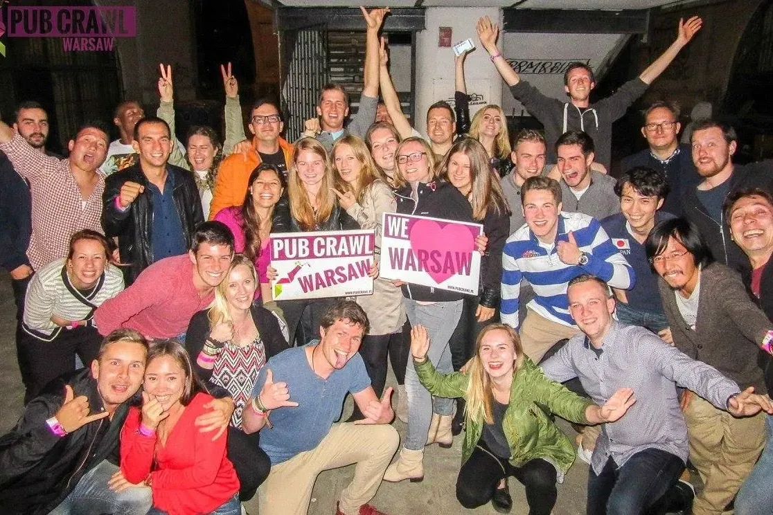 Eine große Gruppe junger Erwachsener lächelt und posiert mit „Pub Crawl Warsaw“- und „Wir lieben Warschau“-Schildern in st...