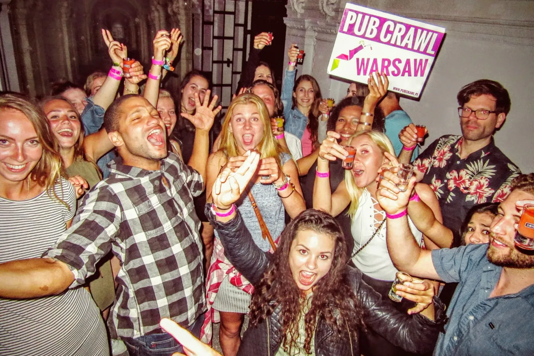 Gruppe junger Erwachsener lachend und lächelnd, mit Schnapsgläsern, einer hält ein 'PUB CRAWL WARSAW' Schild.