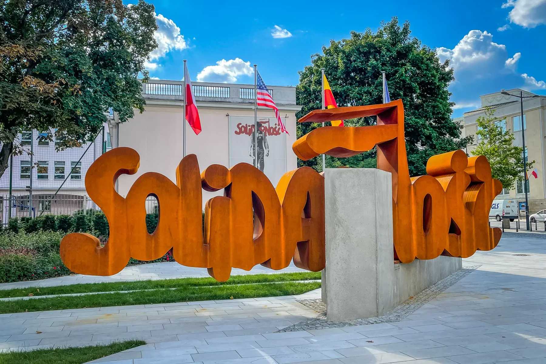 Rust-colored sculpture spelling 'Solidarność' in Warsaw. Polish and US flags, trees, and a white building are visible behi...
