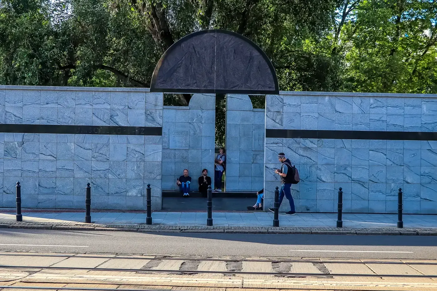 Grey marble memorial wall with a dark semi-circular arch, people standing and sitting, tram tracks and street in foreground.
