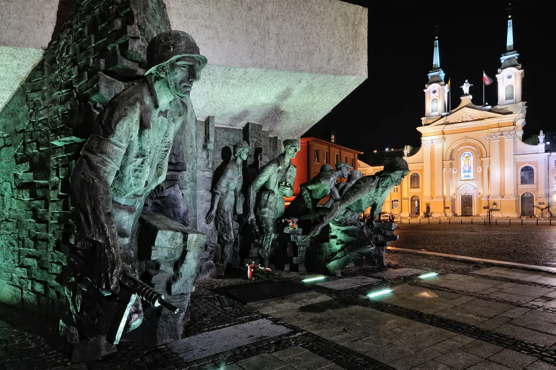 Bronzemonument mit Soldaten des Warschauer Aufstands, nachts grün beleuchtet, im Hintergrund eine hell erleuchtete Kirche.