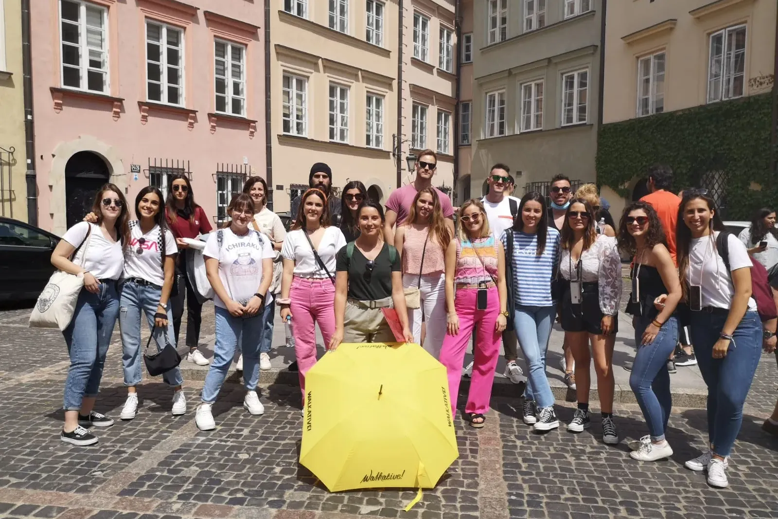 Un grupo de jóvenes posa en una calle empedrada con un paraguas amarillo de Walkative. Edificios históricos coloridos al f...