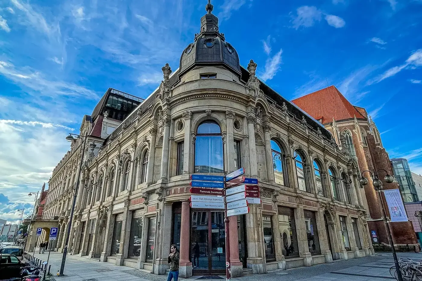 Ornate historic building with a domed corner tower and large windows on a cobblestone street in Wrocław. A street signpost...