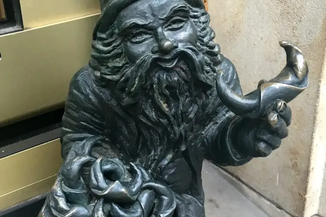 Brązowy posąg krasnala z zaostrzoną czapką i długą brodą, trzymający tacę z splotami i przedmiot w kształcie rogu, przy śc...