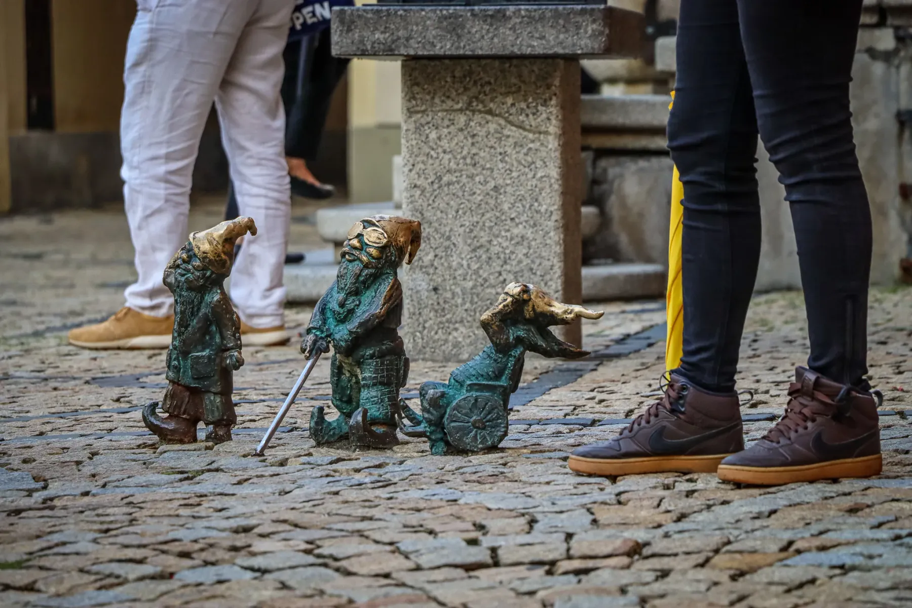 Trzy brązowe figurki krasnali na brukowanej ulicy. Wokół widoczne nogi ludzi. Jeden krasnal trzyma laskę.