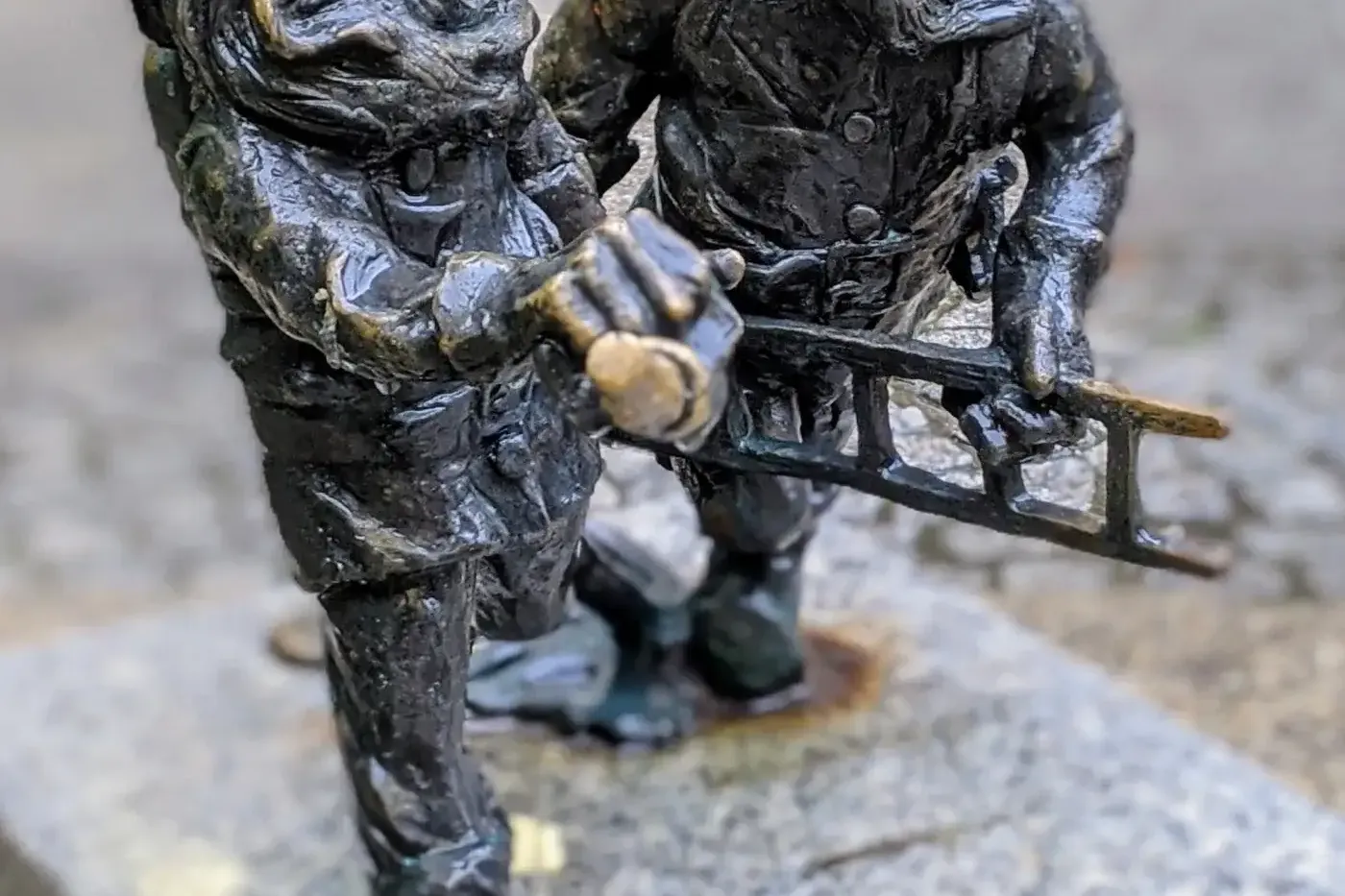Dwie ciemne, brązowe figurki krasnali z Wrocławia w złotych hełmach, jeden z brodą trzymający kilof, drugi drabinę.