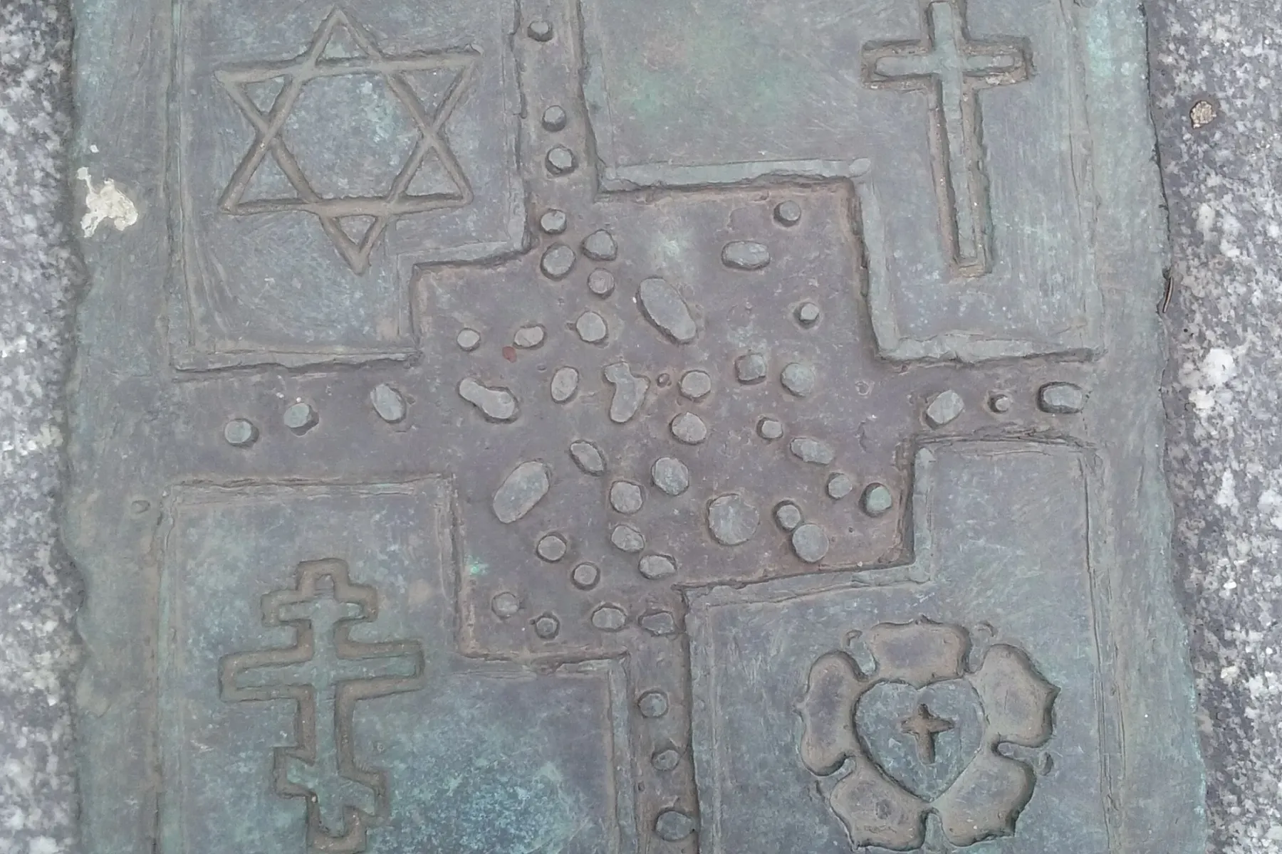 Brązowa tablica w granicie z czterema symbolami religijnymi: Gwiazda Dawida, krzyż łaciński, krzyż prawosławny i róża Lutra.