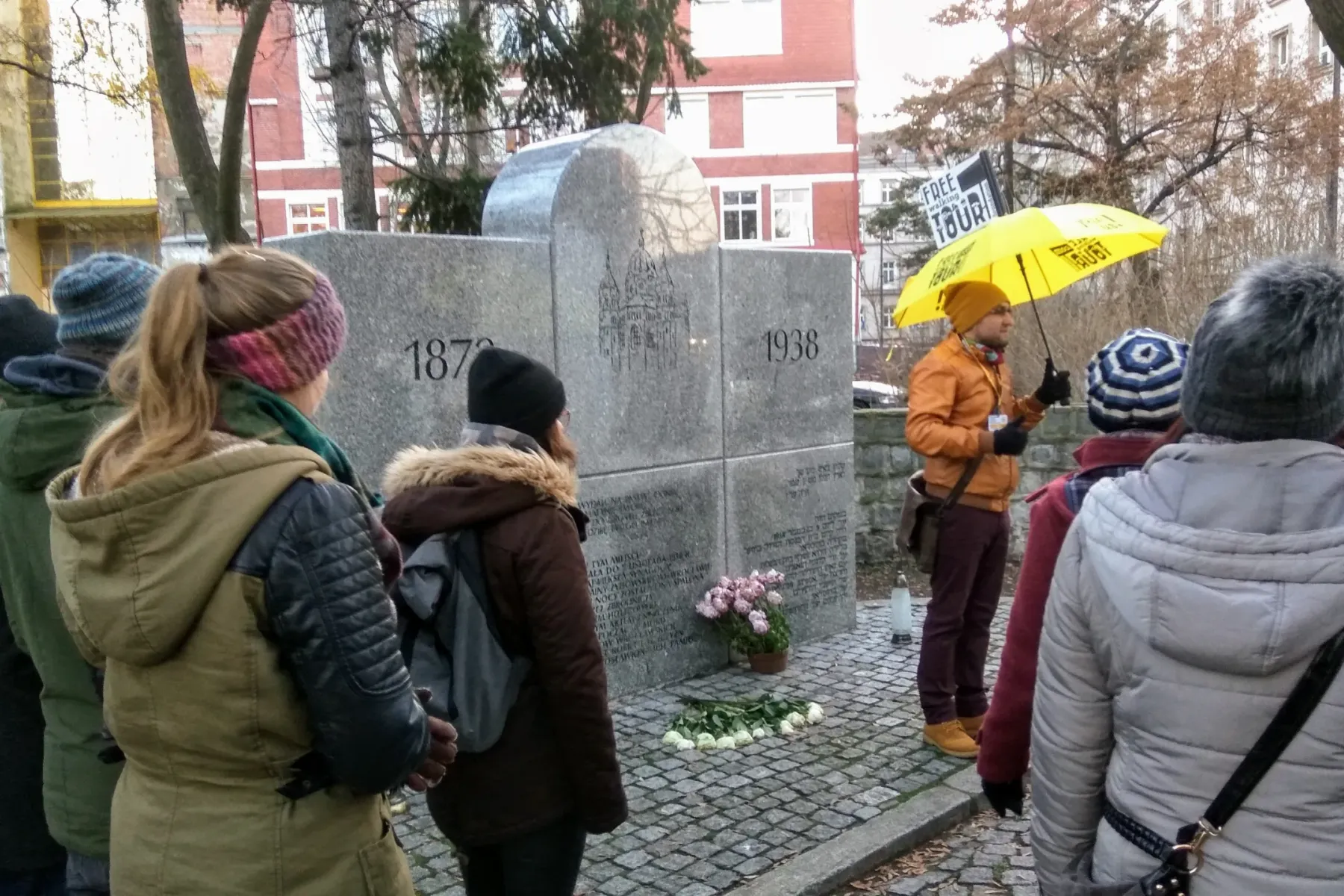 Kamienny pomnik upamiętniający Nową Synagogę z datami 1870 i 1938, w otoczeniu grupy wycieczkowej z przewodnikiem.