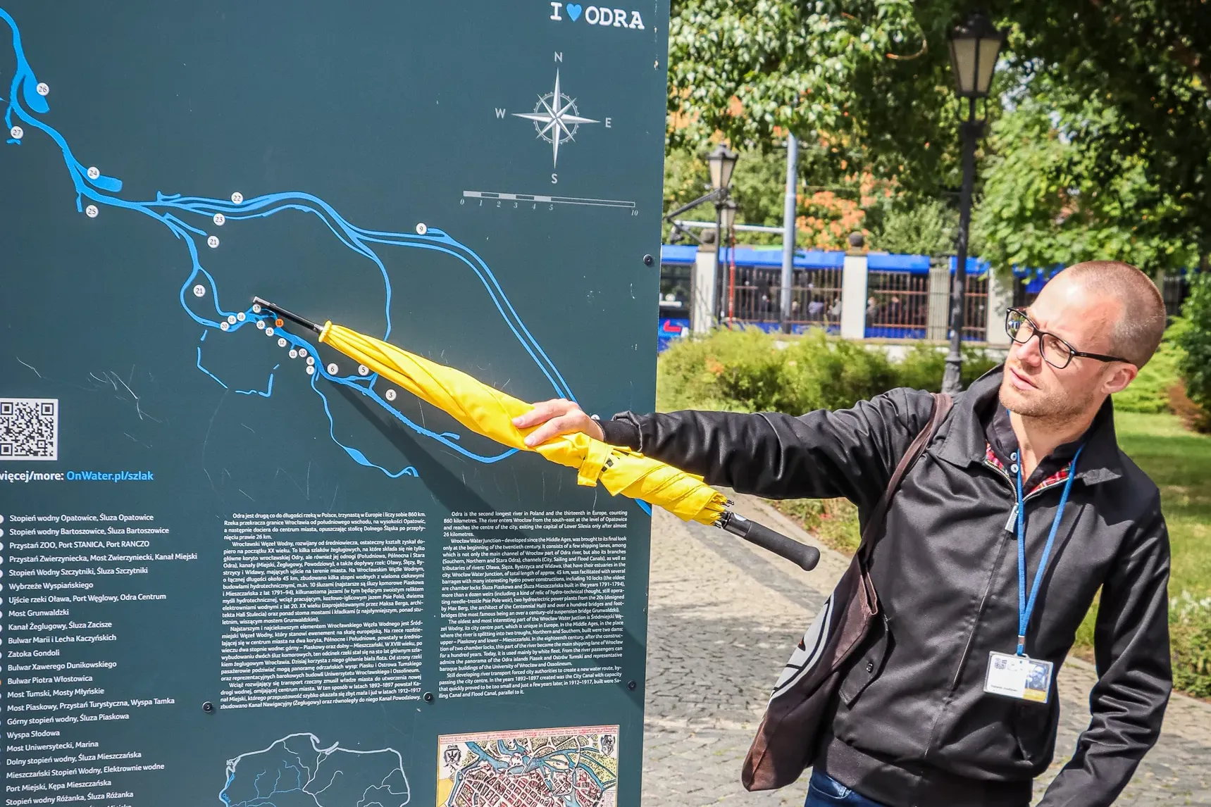 Mężczyzna wskazuje żółtą parasolką tablicę informacyjną z mapą Wrocławskiego Węzła Wodnego w słoneczny dzień.