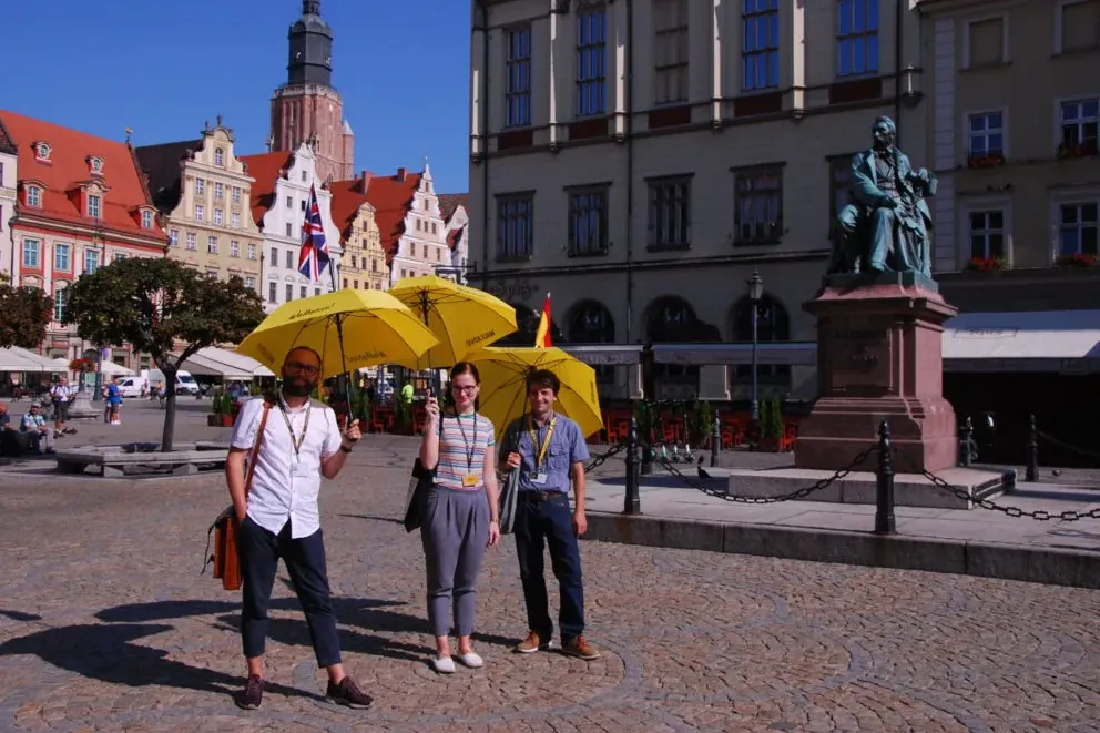 Trzy osoby z żółtymi parasolami na brukowanym rynku, przed historycznymi kamienicami i pomnikiem w słoneczny dzień.