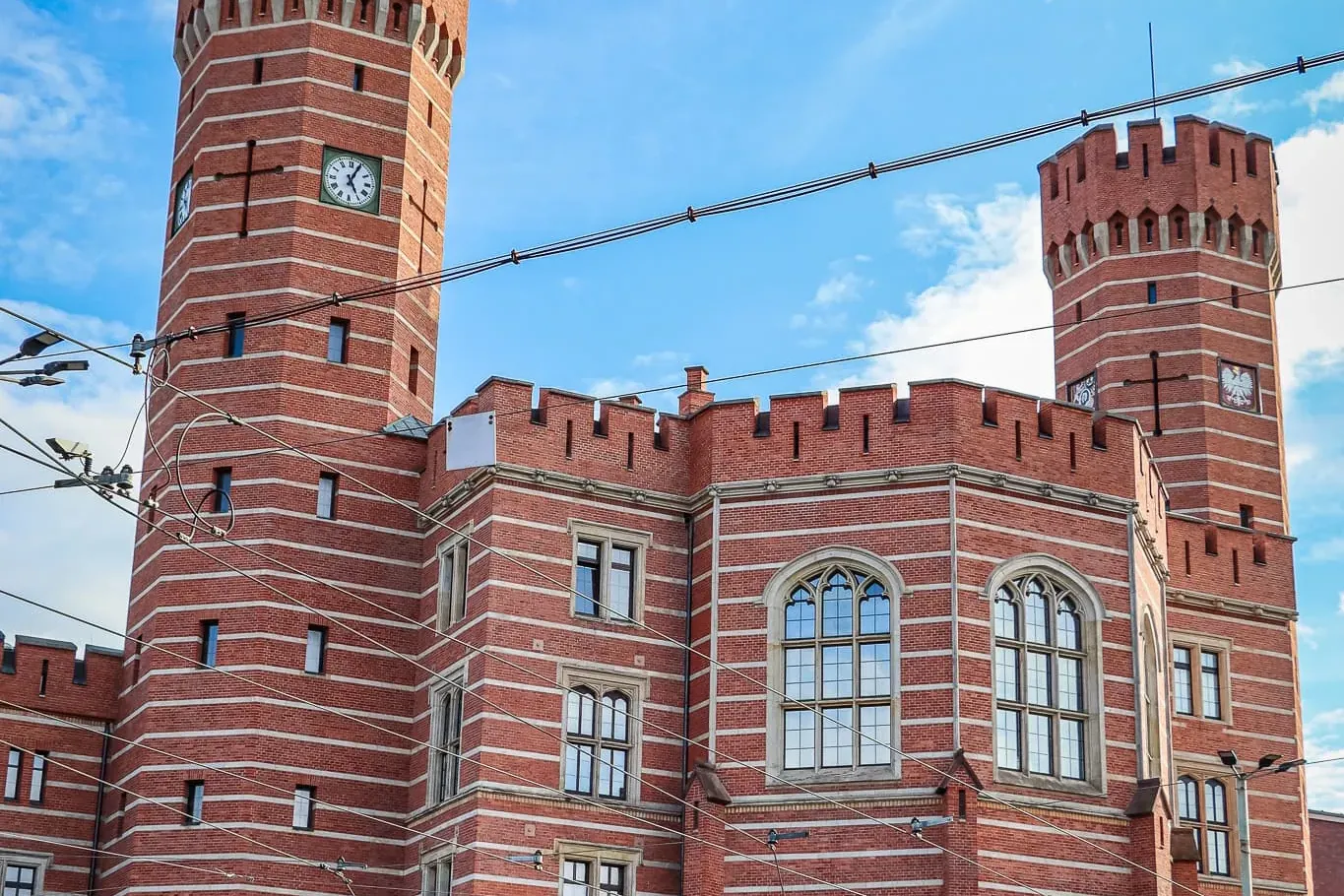 Historyczny budynek z czerwonej cegły z dwiema wieżami zegarowymi, pod błękitnym niebem z chmurami we Wrocławiu.