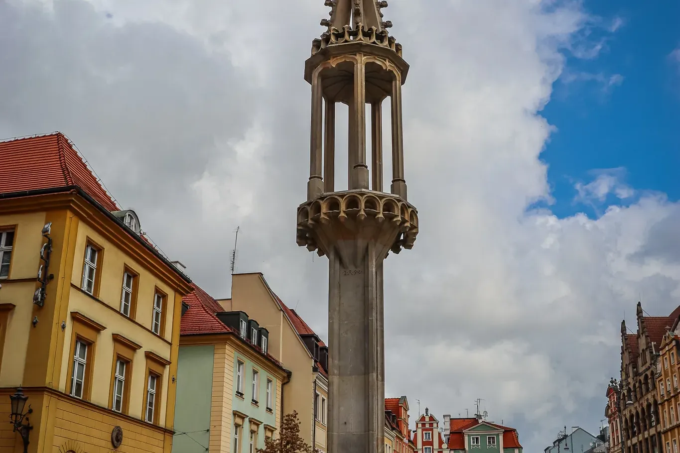 Wysoki kamienny pręgierz z ozdobną iglicą i figurą na szczycie, otoczony kolorowymi zabytkowymi budynkami na rynku.