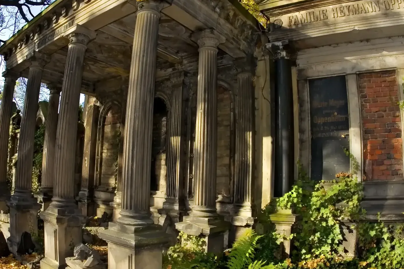 Stone mausoleum with classical columns, intricate stonework, and green ivy, surrounded by fallen yellow autumn leaves unde...