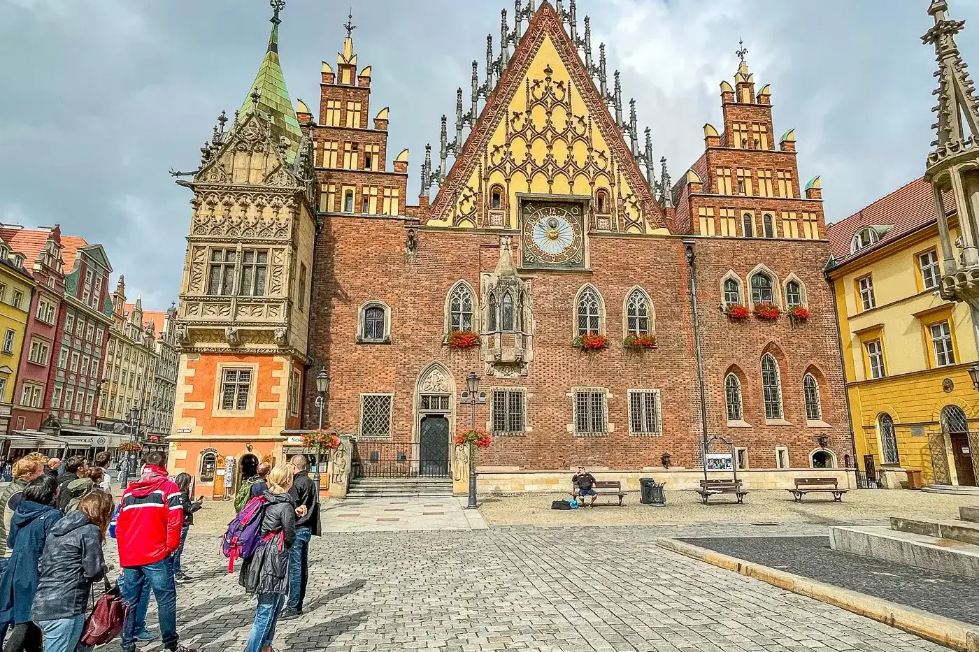 Wrocławski Ratusz, duży gotycki budynek z cegły z wysoką wieżą zegarową, stoi na brukowanym Rynku z grupą ludzi.
