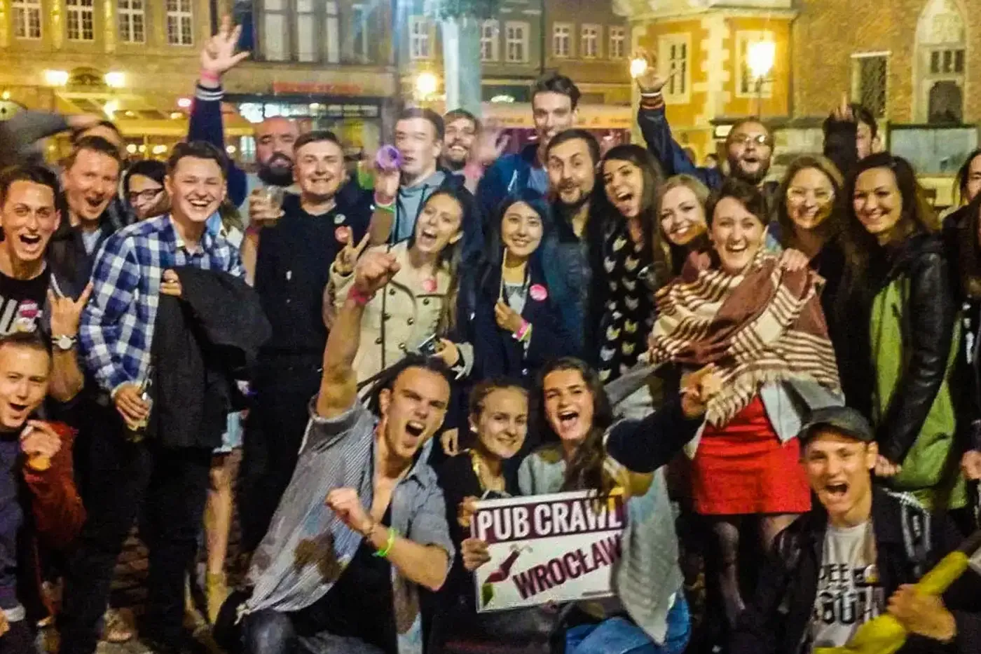 Grupa radosnych osób na nocnym pub crawlu we Wrocławiu, w tle zabytkowe kamienice. Jedna osoba trzyma tabliczkę "Pub Crawl...