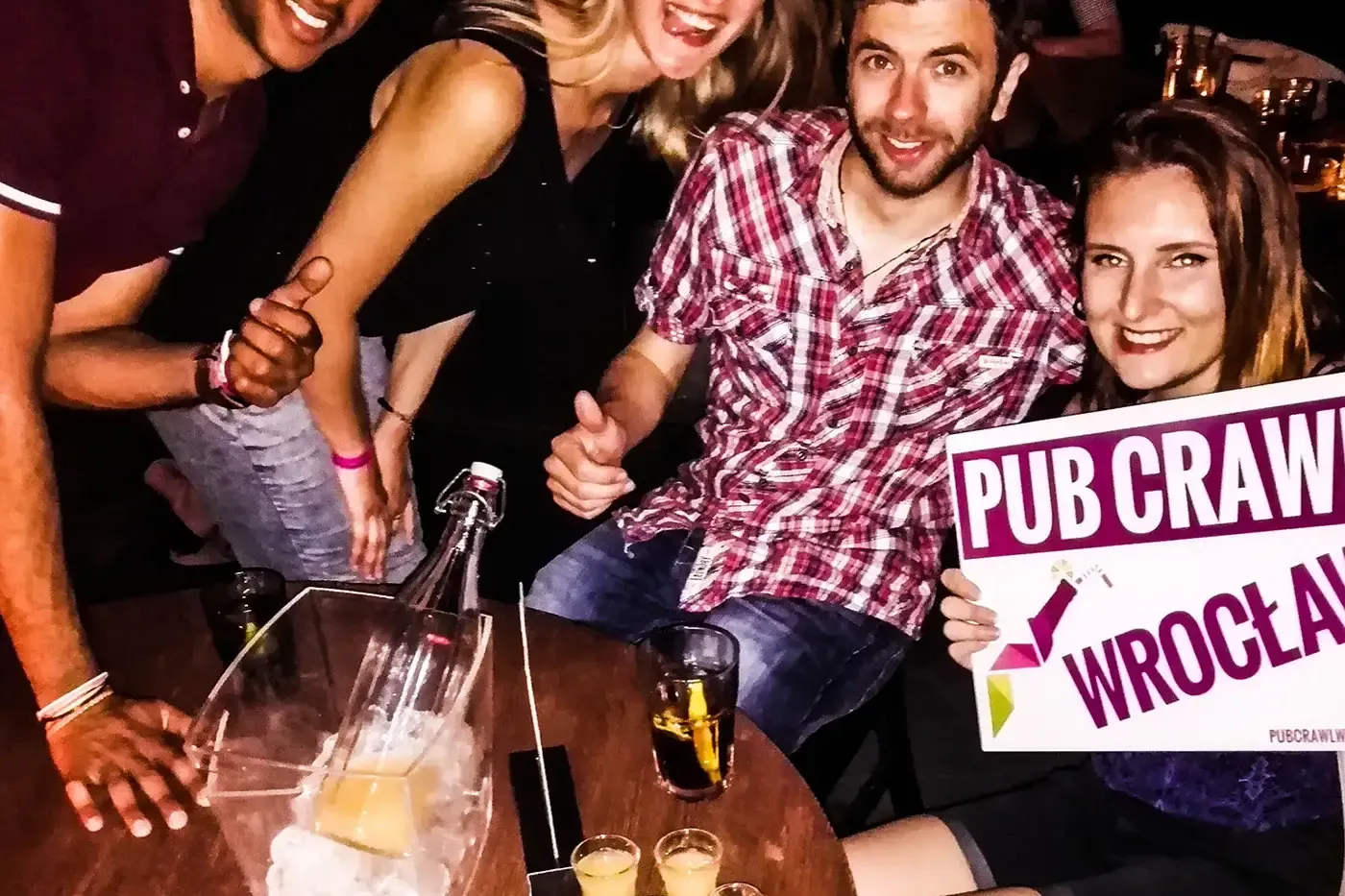 Cztery uśmiechnięte osoby przy stole z napojami i szotami, jedna trzyma znak 'PUB CRAWL WROCŁAW'.