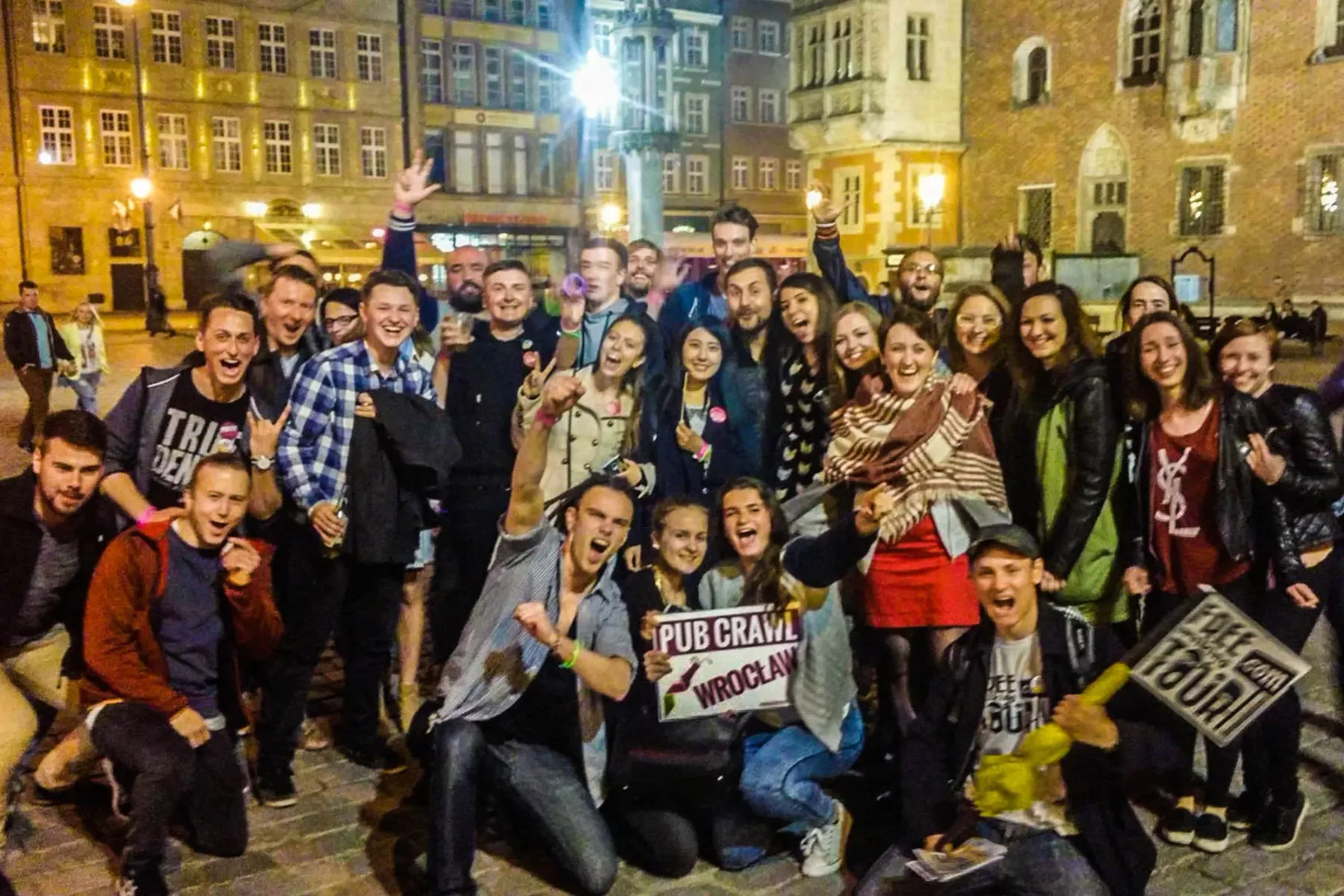 Duża grupa młodych dorosłych uśmiechniętych i pozujących do zdjęcia na ulicy nocą. Jedna osoba trzyma tablicę 'Pub Crawl W...