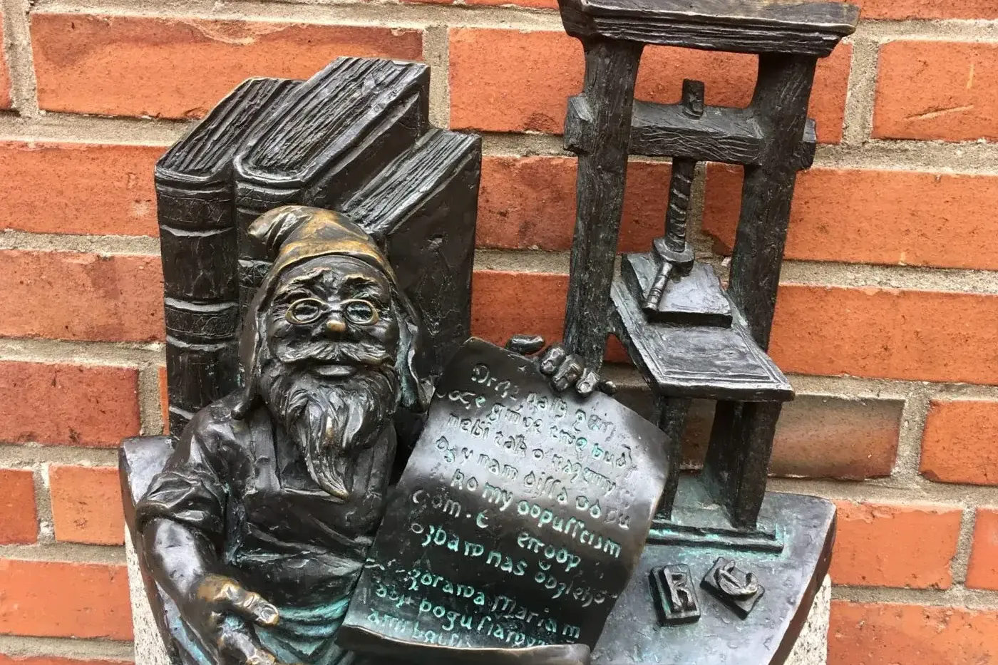 Brązowa statuetka krasnala w okularach, trzymającego dokument, obok książek i prasy drukarskiej, na tle czerwonej ceglanej...