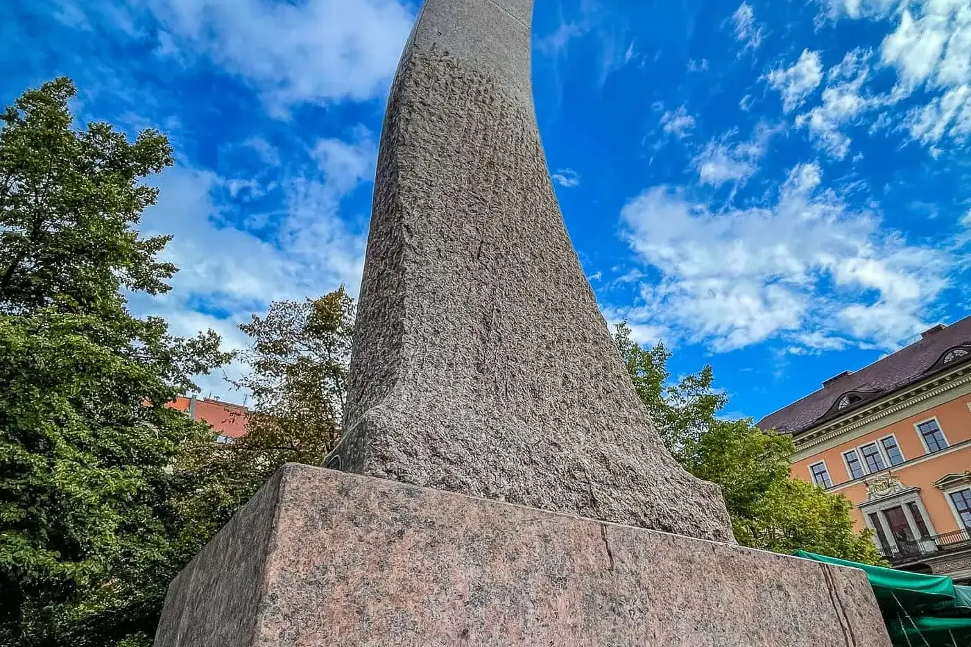 Wysoki, spiralny abstrakcyjny pomnik kamienny o chropowatej fakturze, na granitowej podstawie, pod błękitnym niebem z chmu...